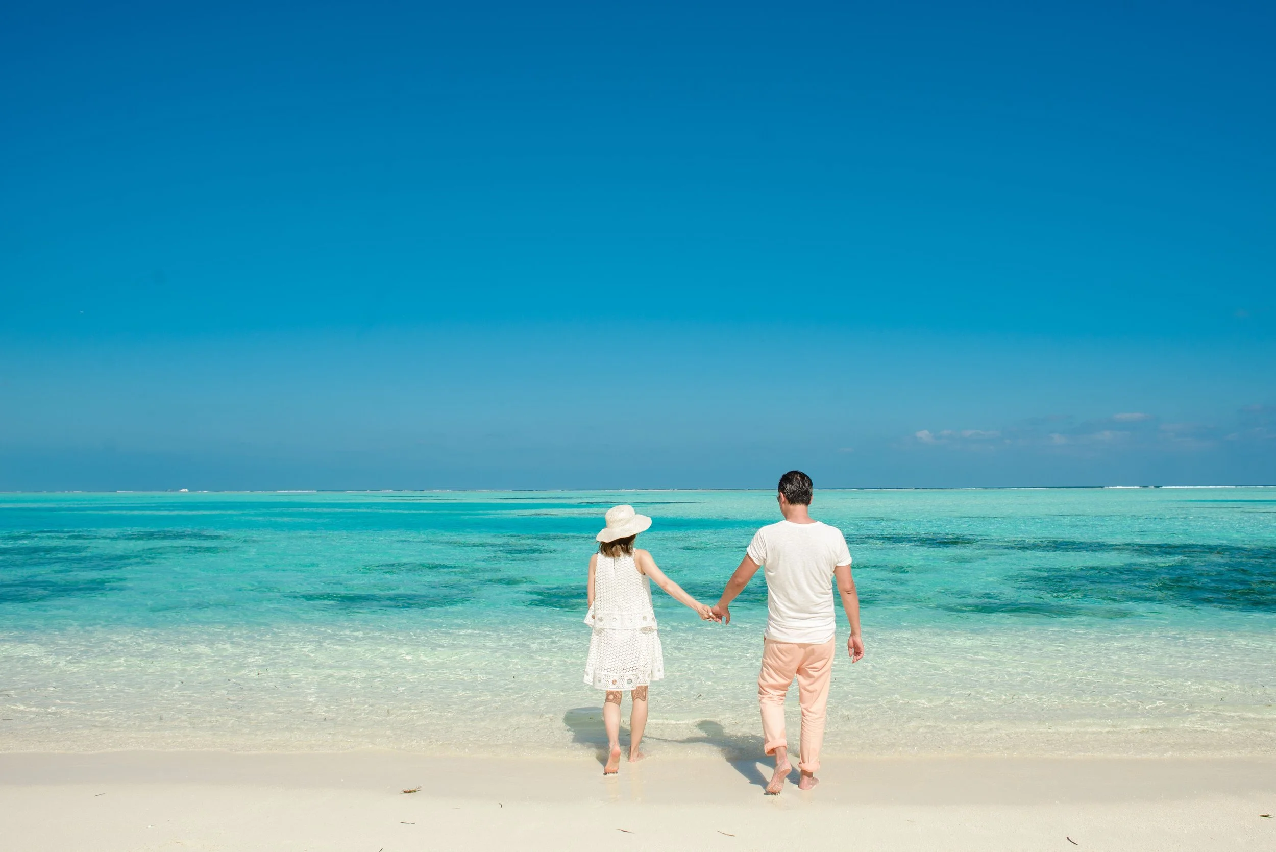 A couple holding hands walking towards the ocean on a sandy beach with clear turquoise water and blue sky.