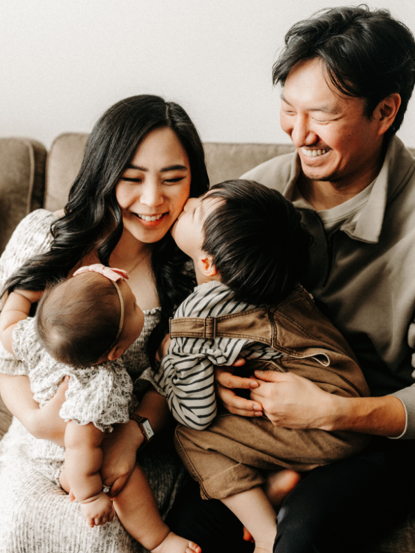 Family of four sitting on a couch, smiling, with two young children, a boy and a girl, sharing a close moment in a cozy living room.
