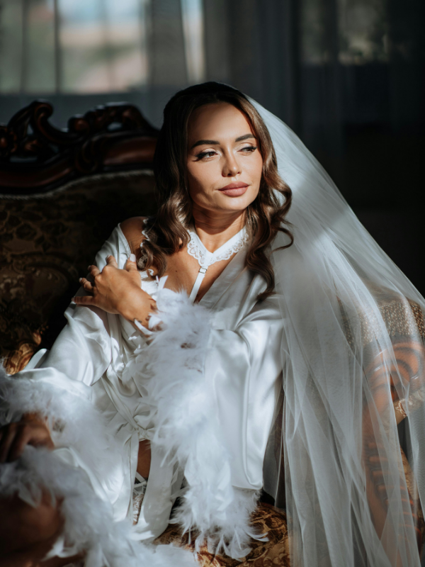 A bride in a white satin robe and veil sitting on an ornate vintage sofa, gazing thoughtfully out of a window.