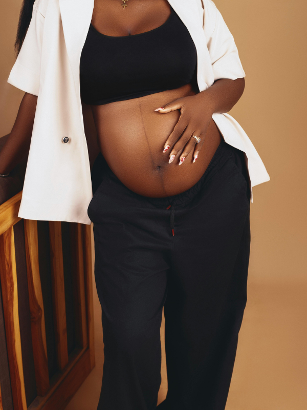 A pregnant woman with medium skin tone wearing a black top, open white blazer, and black pants, standing indoors with one hand on her belly and the other hand resting on a wooden piece of furniture, against a beige background.