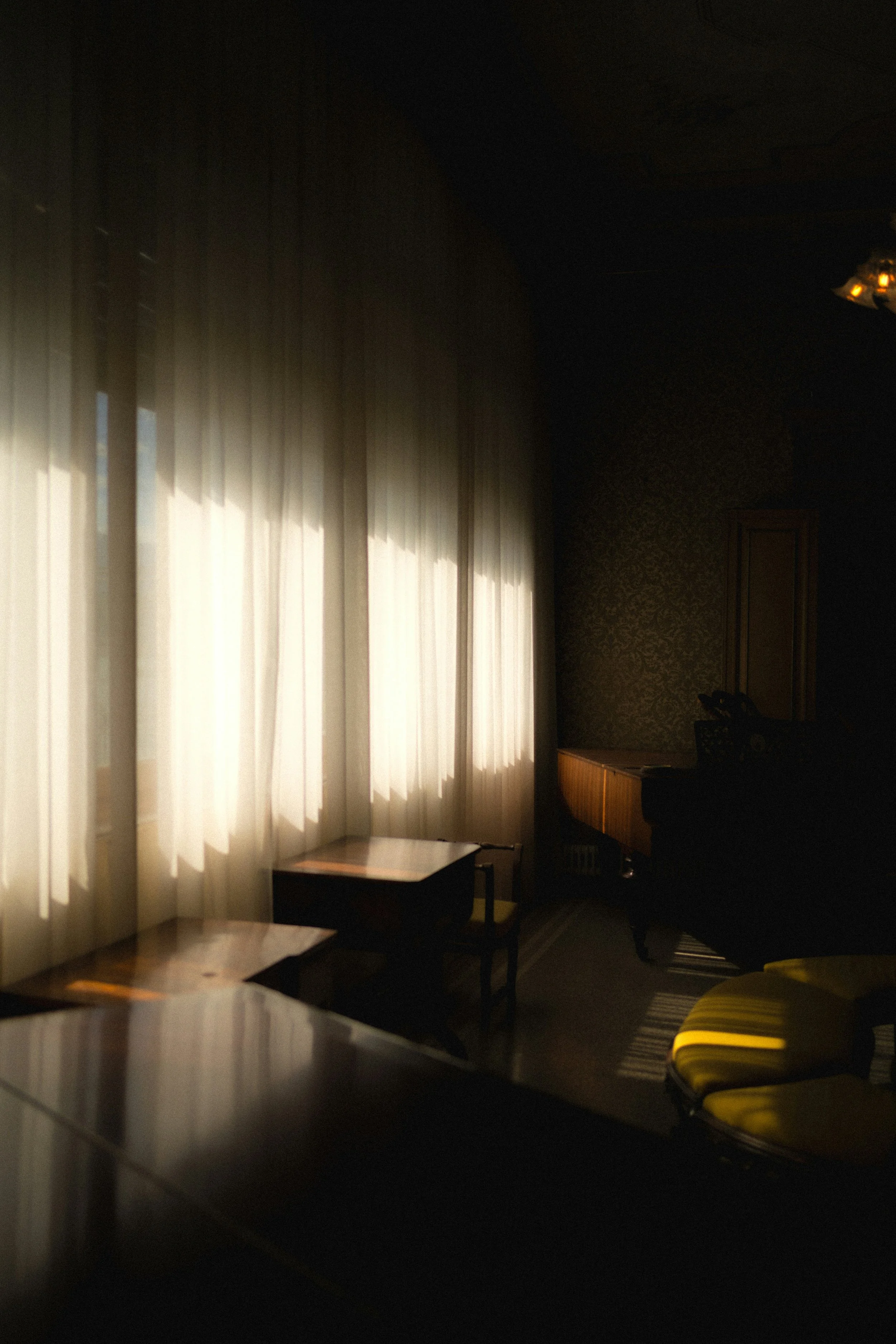 A dimly lit room with beige curtains on the left side, sunlight filtering through. There is a small table and a yellow chair with black legs in the foreground. In the background, there is a wooden piano and some chairs, with dark walls and a patterned wallpaper. The room appears to be a living or sitting area.