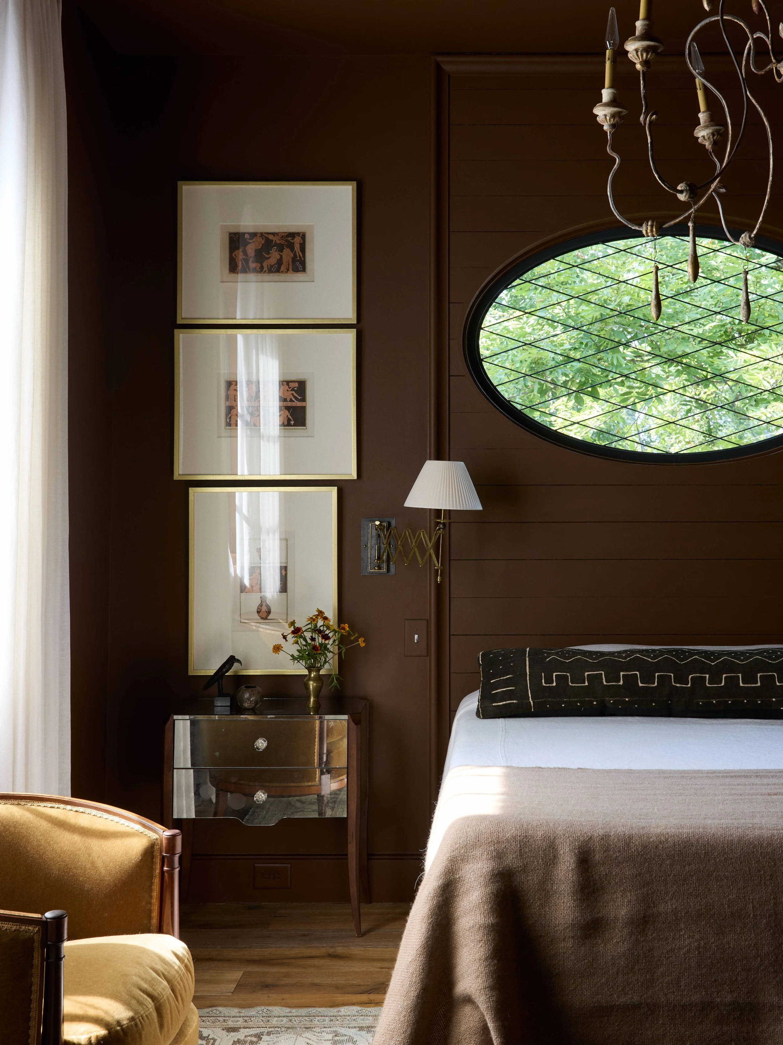 An interior bedroom scene with a bed partially visible on the right, a side table with a lamp, and framed artwork on a brown wall. A decorative chandelier hangs from the ceiling, and a round window with greenery outside is also in view.