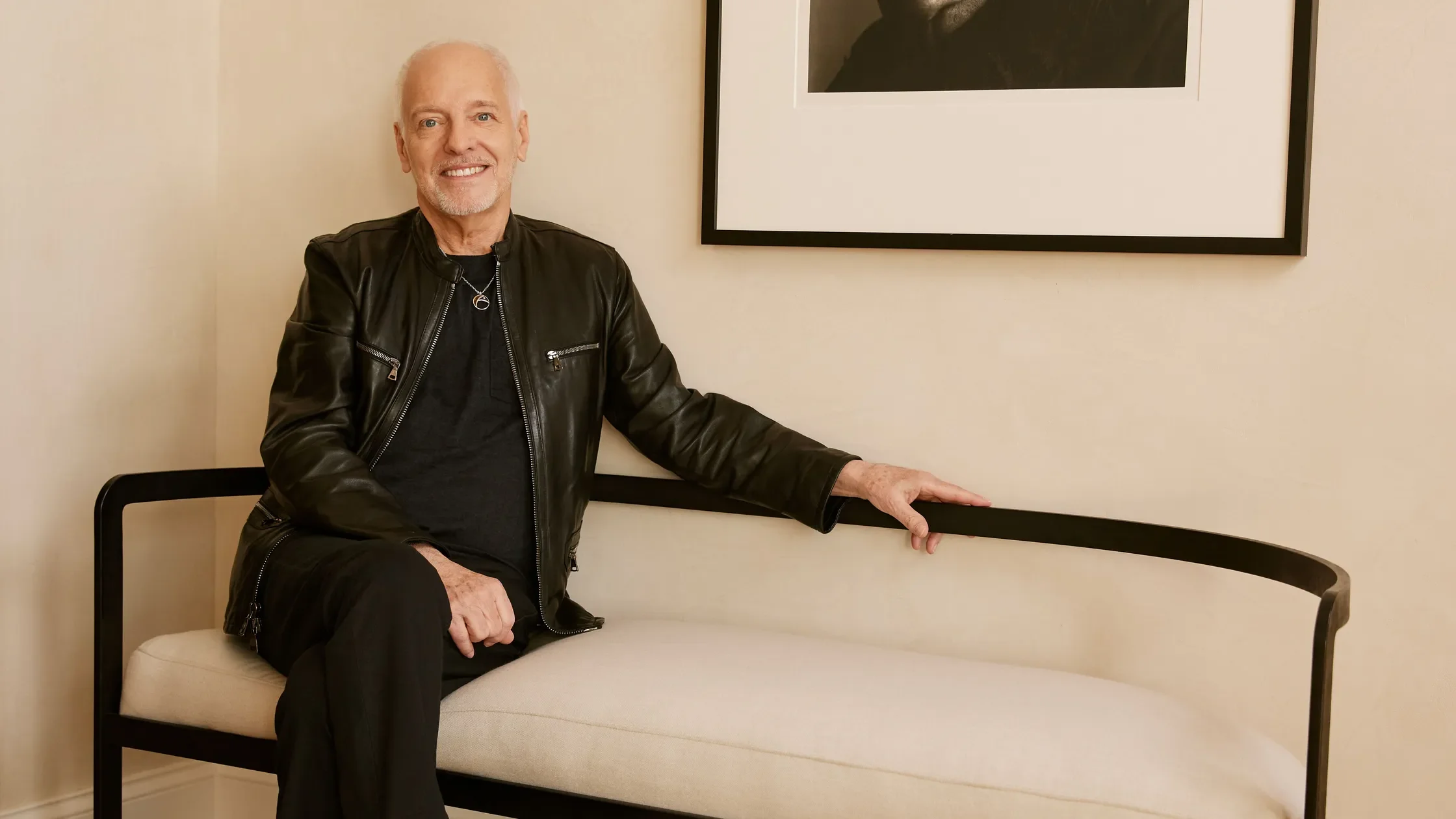 An elderly man with gray hair and a beard, smiling, wearing a black leather jacket and a black shirt, sitting on a white couch in a room with beige walls and a framed black-and-white picture hanging on the wall.