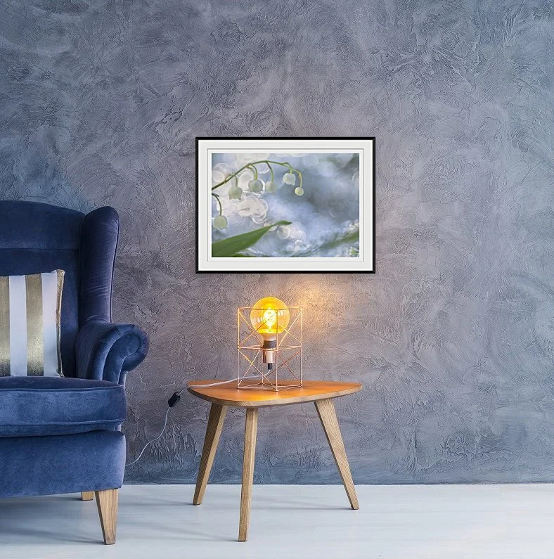 A cozy corner with a blue armchair, a round wooden side table with a decorative lamp, and a framed photograph of white flowers on a textured gray wall.