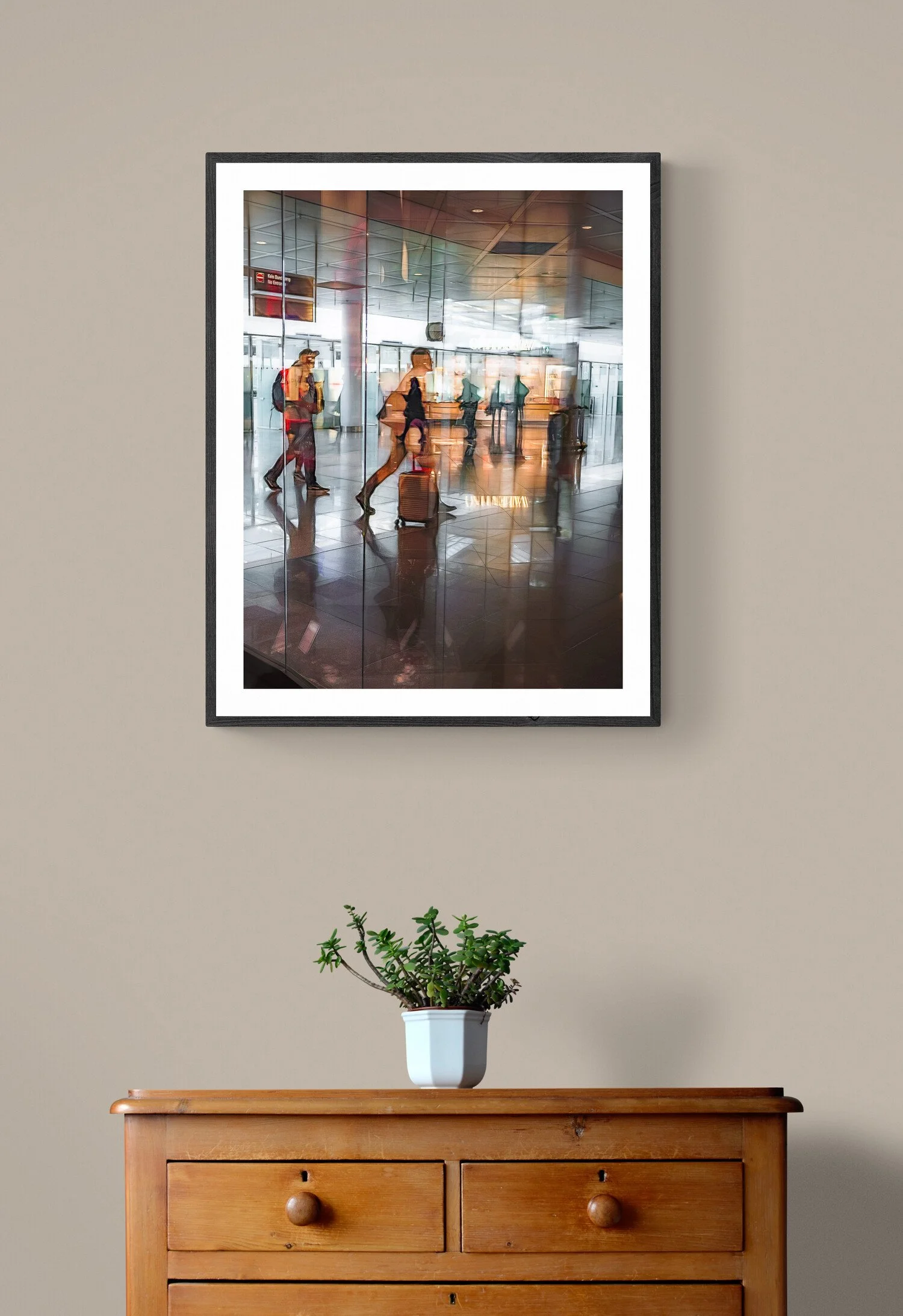 A wooden dresser with a potted green plant on top, mounted on a beige wall, with a framed photograph of travelers in an airport reflected in the glass.