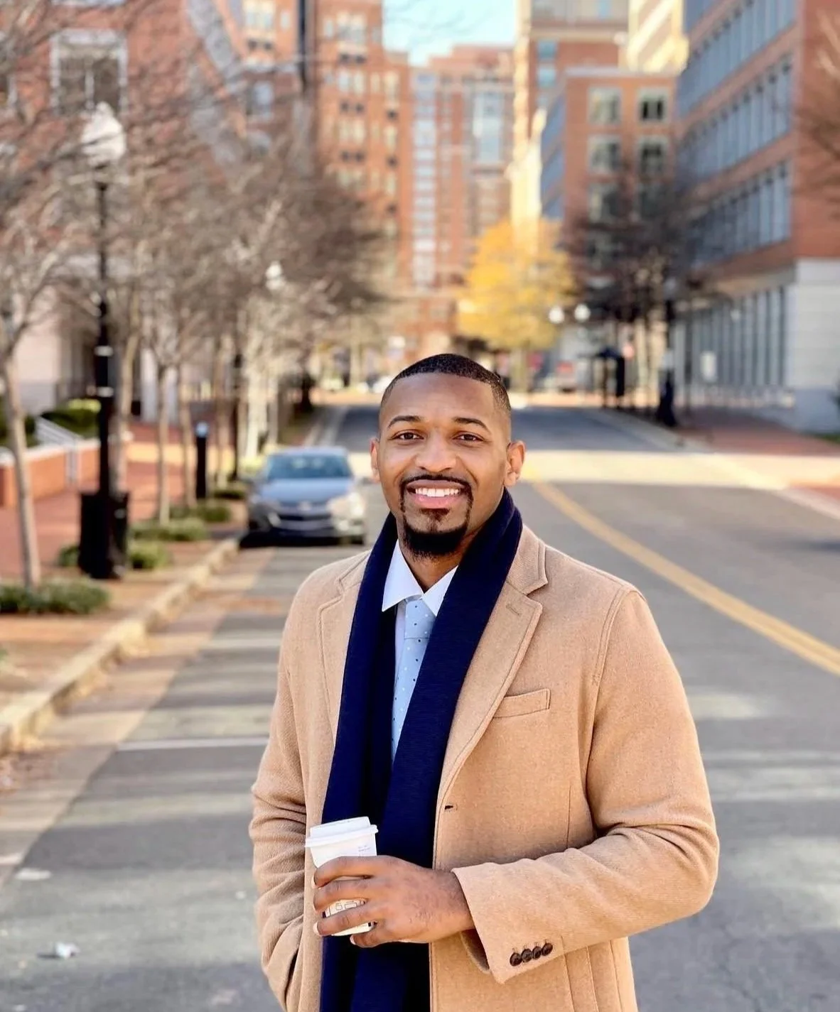 A smiling man in a tan coat and dark scarf holding a coffee cup standing on a city sidewalk with trees and buildings in the background.