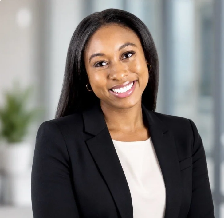 Smiling professional woman with long black hair, wearing a black blazer and white blouse, standing in an office with blurred background.