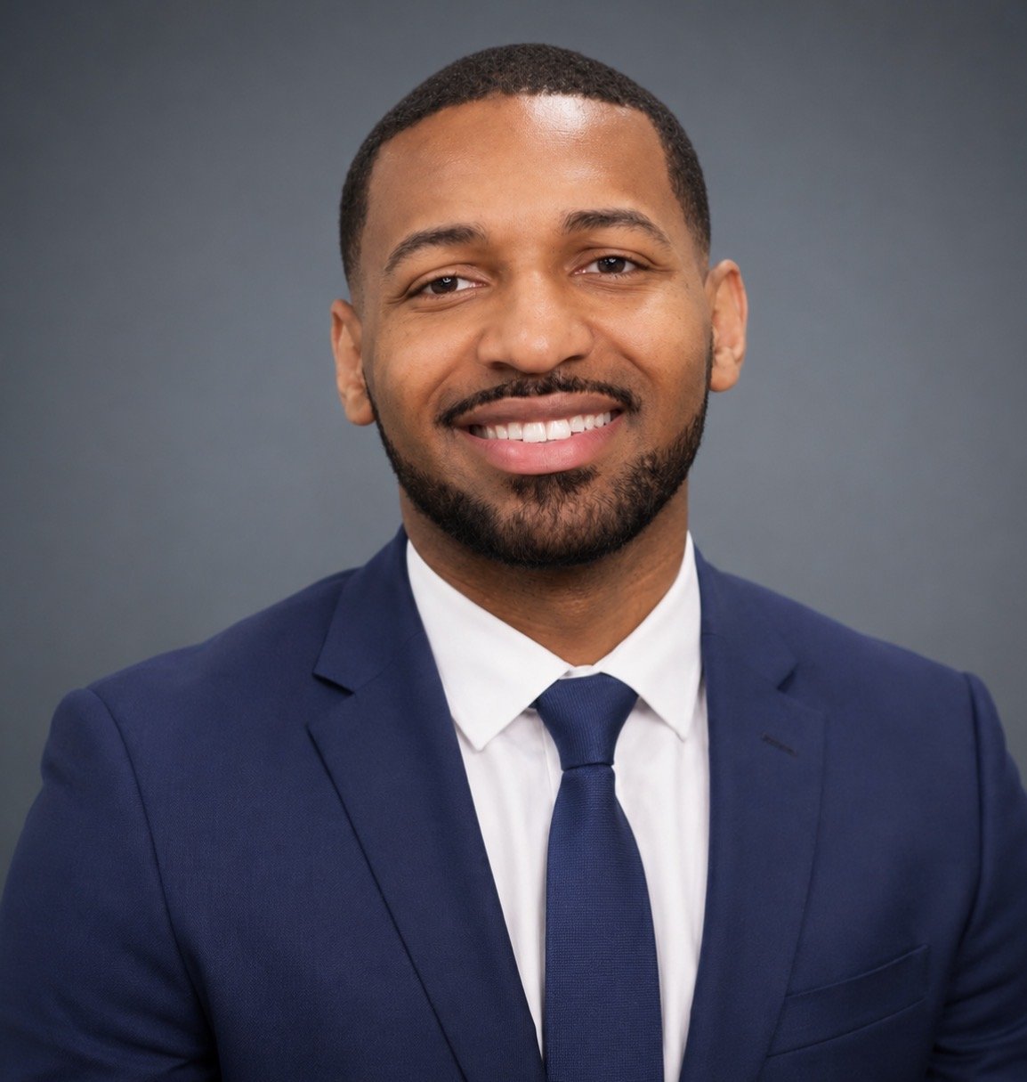 Headshot of a man in a navy suit, white shirt, and navy tie, smiling against a gray background.
