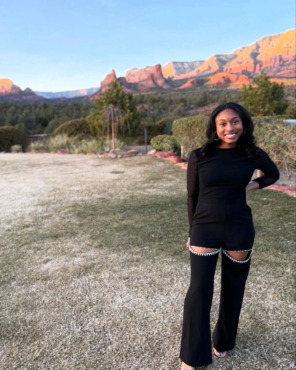 A woman in black clothing stands outdoors with a smile, in front of a scenic mountainous landscape with rocky formations and trees.