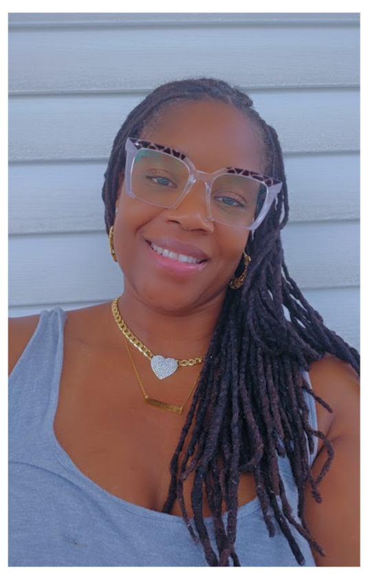 A woman with long twisted hair wearing sunglasses, a gray tank top, and gold jewelry smiling in front of a light gray wooden wall.