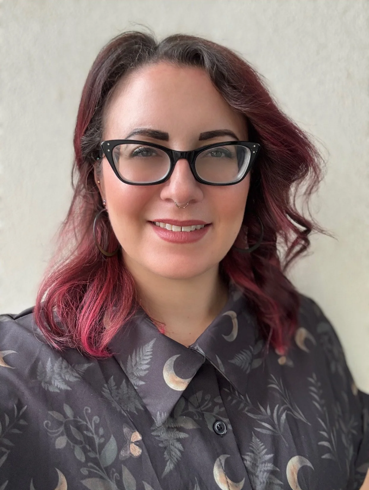 A woman with glasses, dark hair with pink tips, smiling in front of a plain wall, wearing a shirt with moon and star patterns.