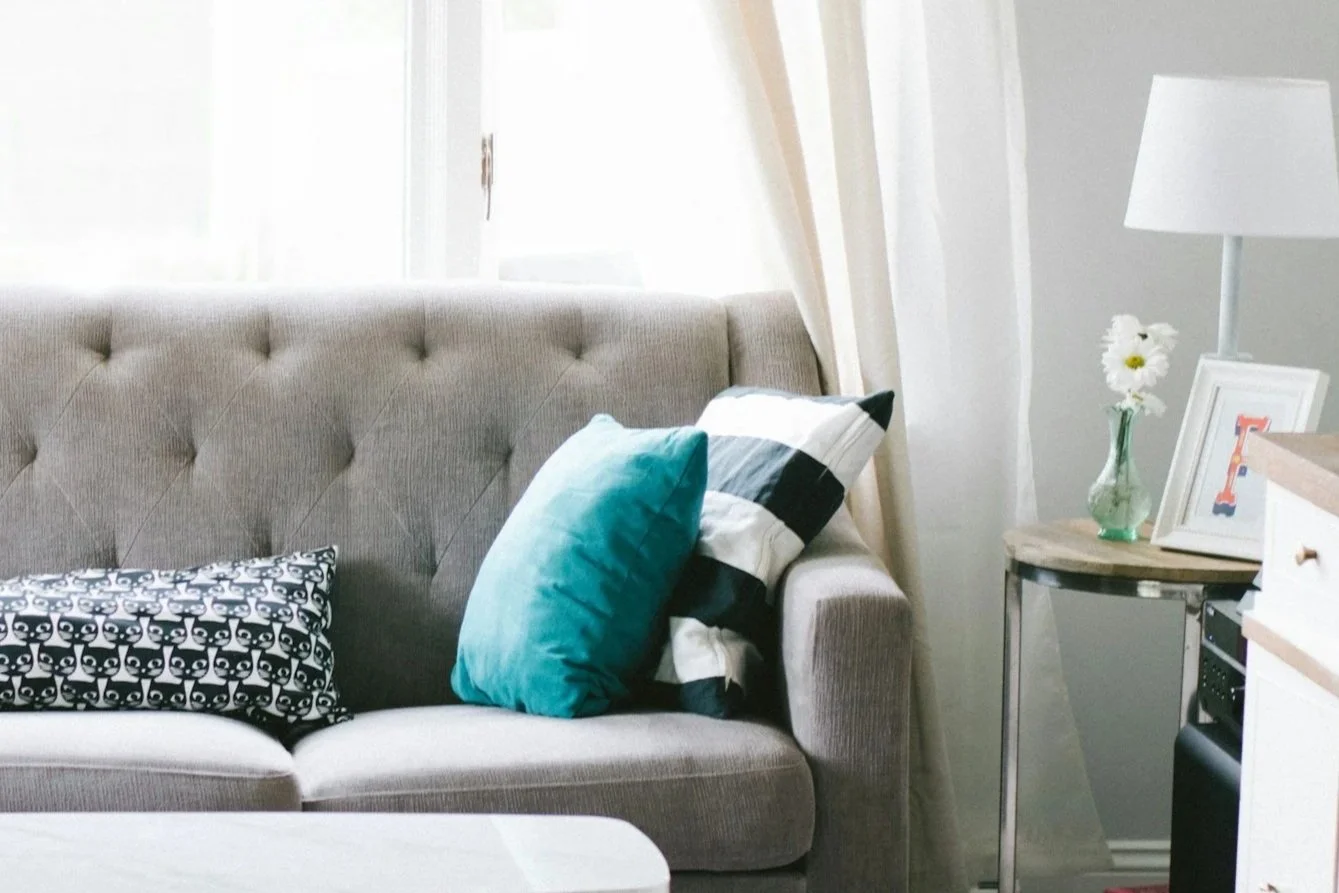 Living room with beige tufted sofa, turquoise and black-and-white pillows, side table with a vase of white flowers, a framed picture, and a modern white lamp.
