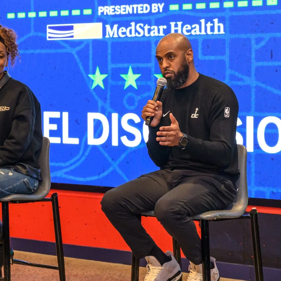 A man sitting on a chair holding a microphone during a panel discussion, with a large screen behind him displaying the MedStar Health logo and text in Spanish, including three green stars.