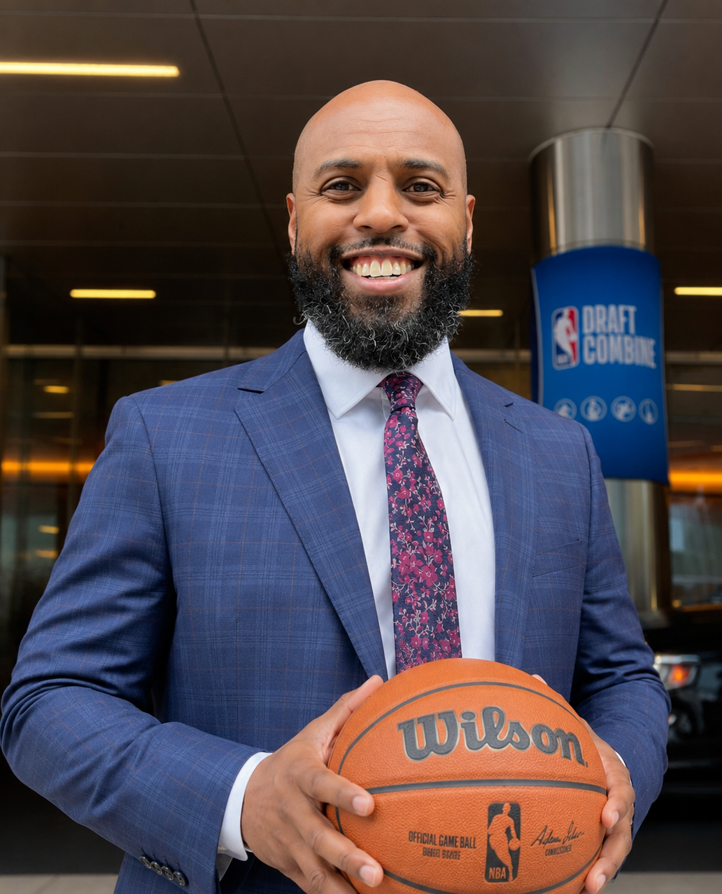 A smiling man in a blue suit holds a Wilson NBA basketball outside a building with a NBA Draft Combine sign in the background.