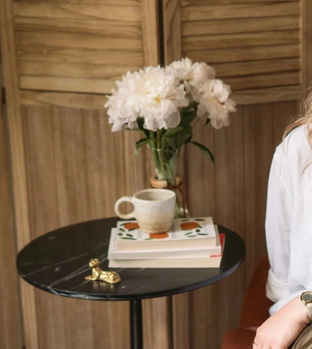 A small black round table with a white ceramic mug, a stack of books, a small gold decorative animal figure, and a glass vase with white peony flowers against a wooden wall.