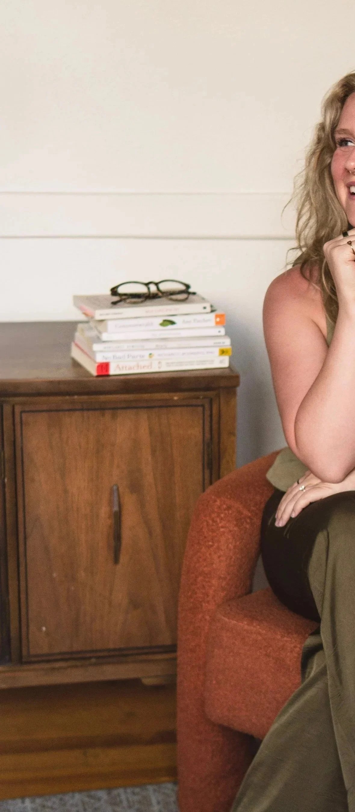 A woman with curly blonde hair smiling, sitting on an armchair, with a stack of books and reading glasses on a wooden cabinet beside her.
