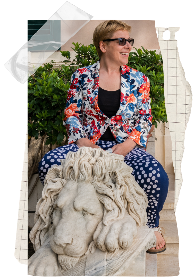 Winter Miller wearing sunglasses, a floral blazer, and polka-dot pants, sitting on a step beside a stone lion statue, and smiling while looking to her right with greenery behind her.