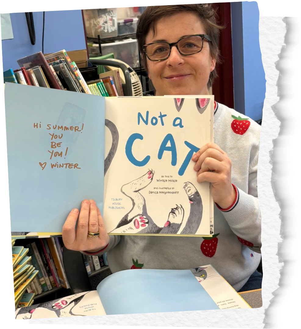 Winter Miller holds up a children's book titled 'Not a Cat' in a library. Winter is wearing a grey sweater with strawberry patches and is smiling. The background has bookshelves and a blue wall.