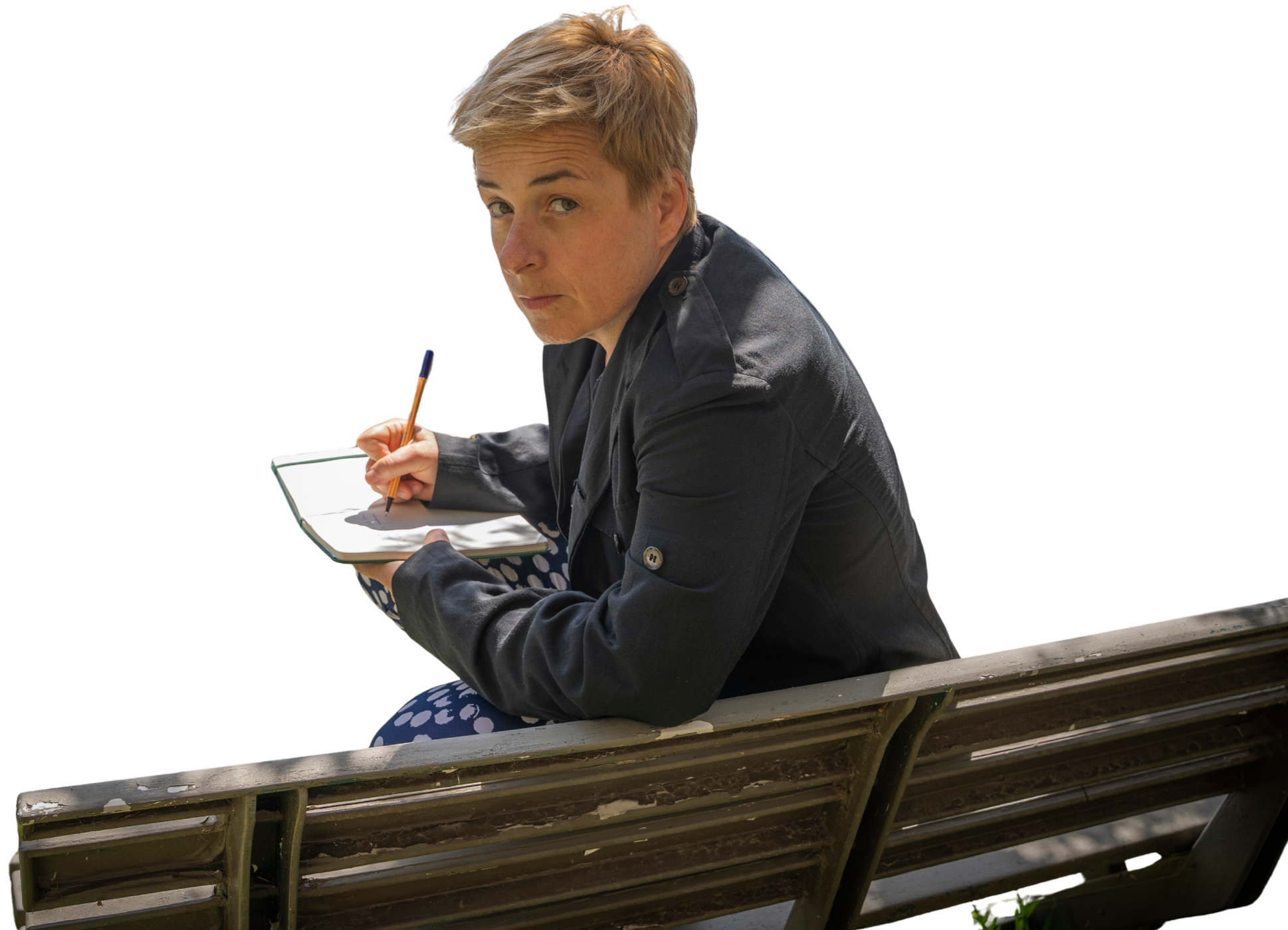 Winter Miller sitting on a park bench, writing in a notebook, wearing a dark jacket and patterned pants.