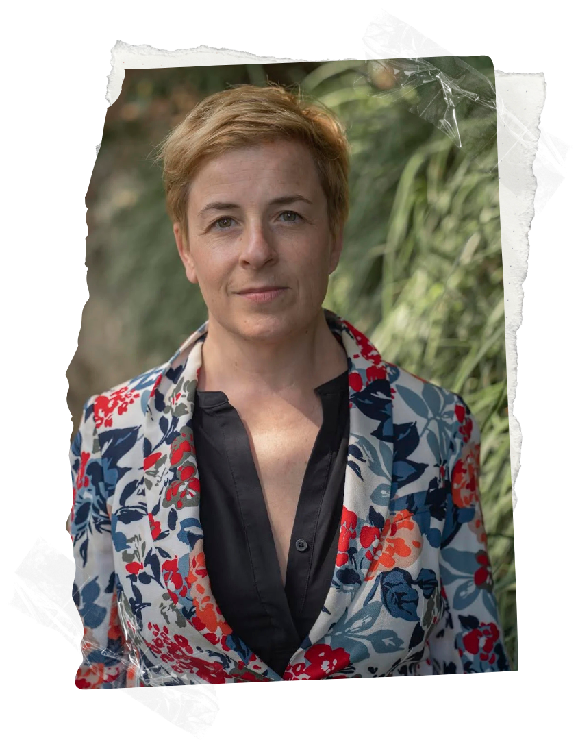 A woman with short, blonde hair and fair skin standing outdoors with green foliage in the background. She is wearing a floral-patterned blazer with red, blue, and gray colors over a black shirt.