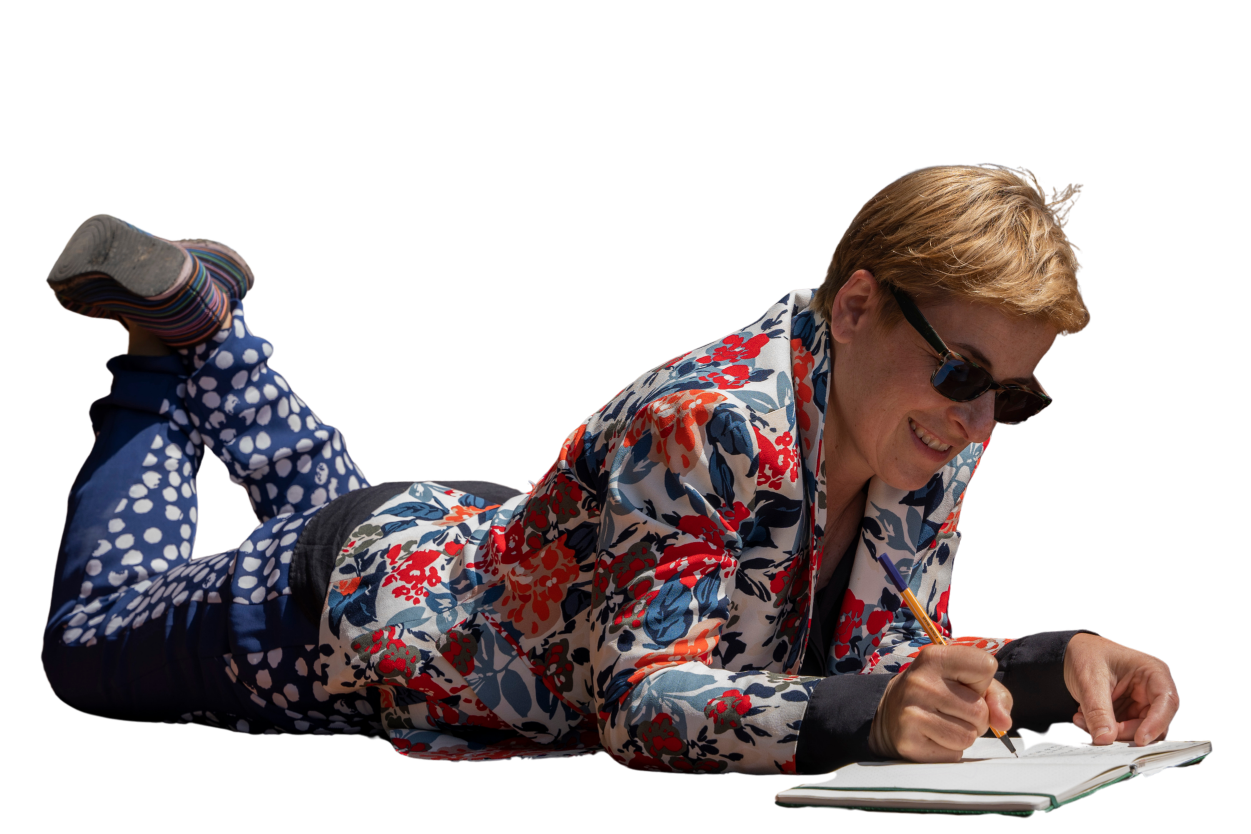 Winter Miller wearing sunglasses and a floral patterned jacket lying on her stomach and writing in a notebook with a pen.
