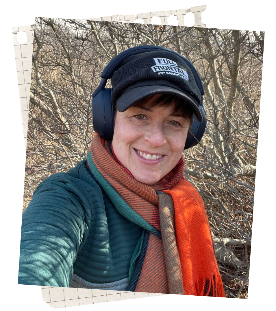A woman taking a selfie outdoors in a wooded area, wearing a dark hat with a patch that says "Full Frontal with Samantha Bee," large headphones, a teal quilted jacket, and an orange scarf, smiling at the camera.