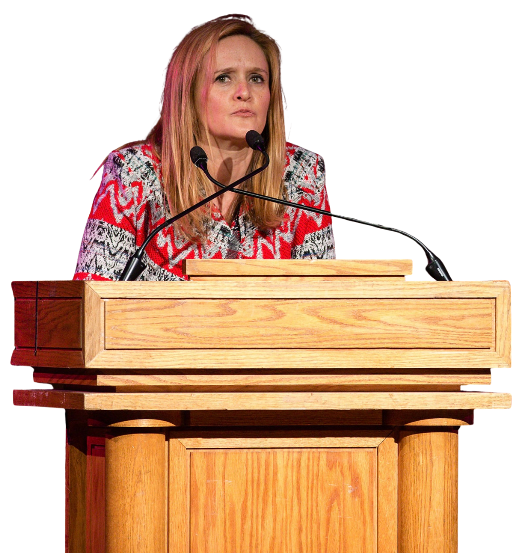 Samantha Bee standing at a wooden podium with two microphones, making a serious facial expression.