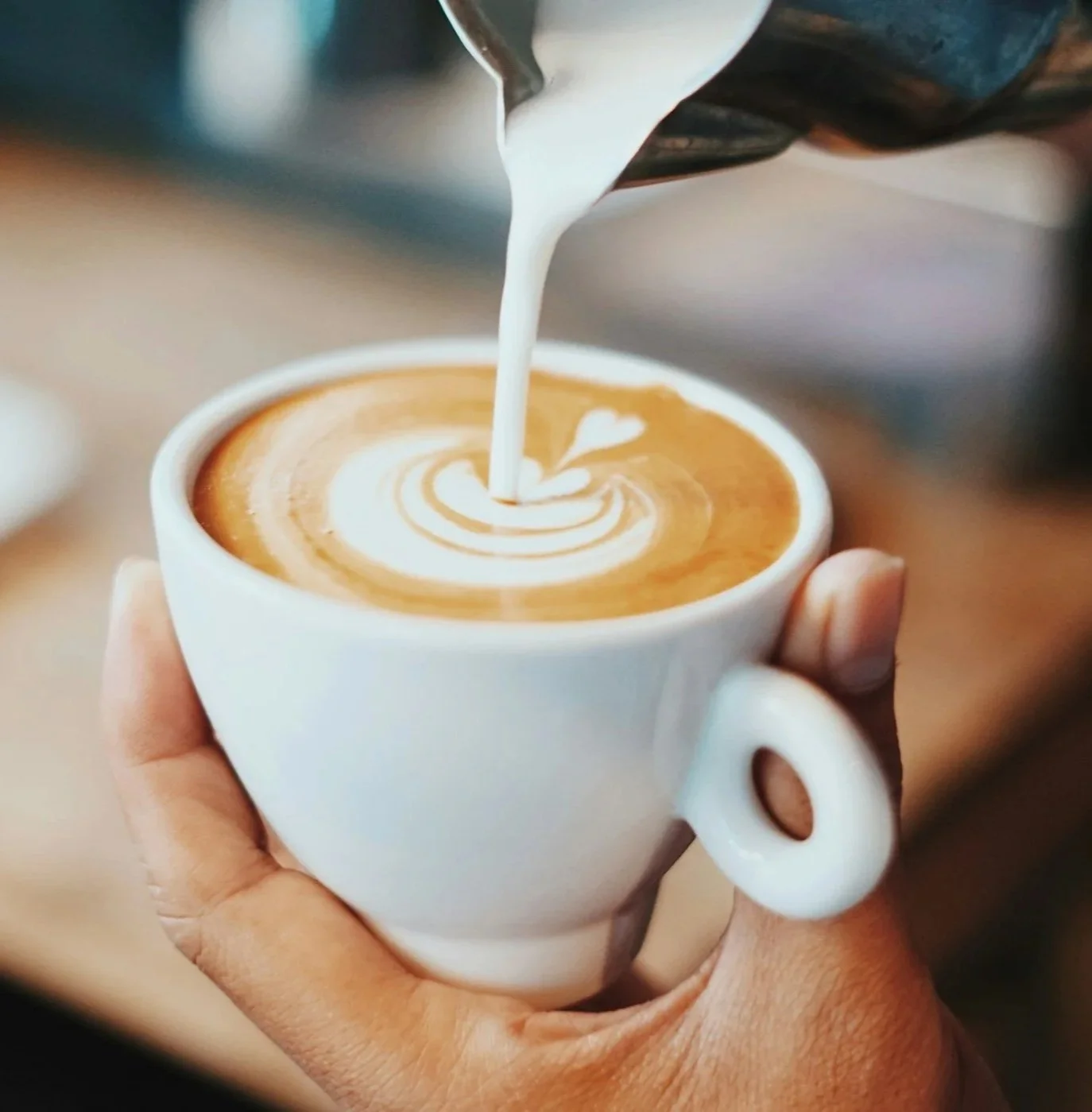 A person holding a white ceramic cup of coffee with latte art, pouring steamed milk to create a heart design.