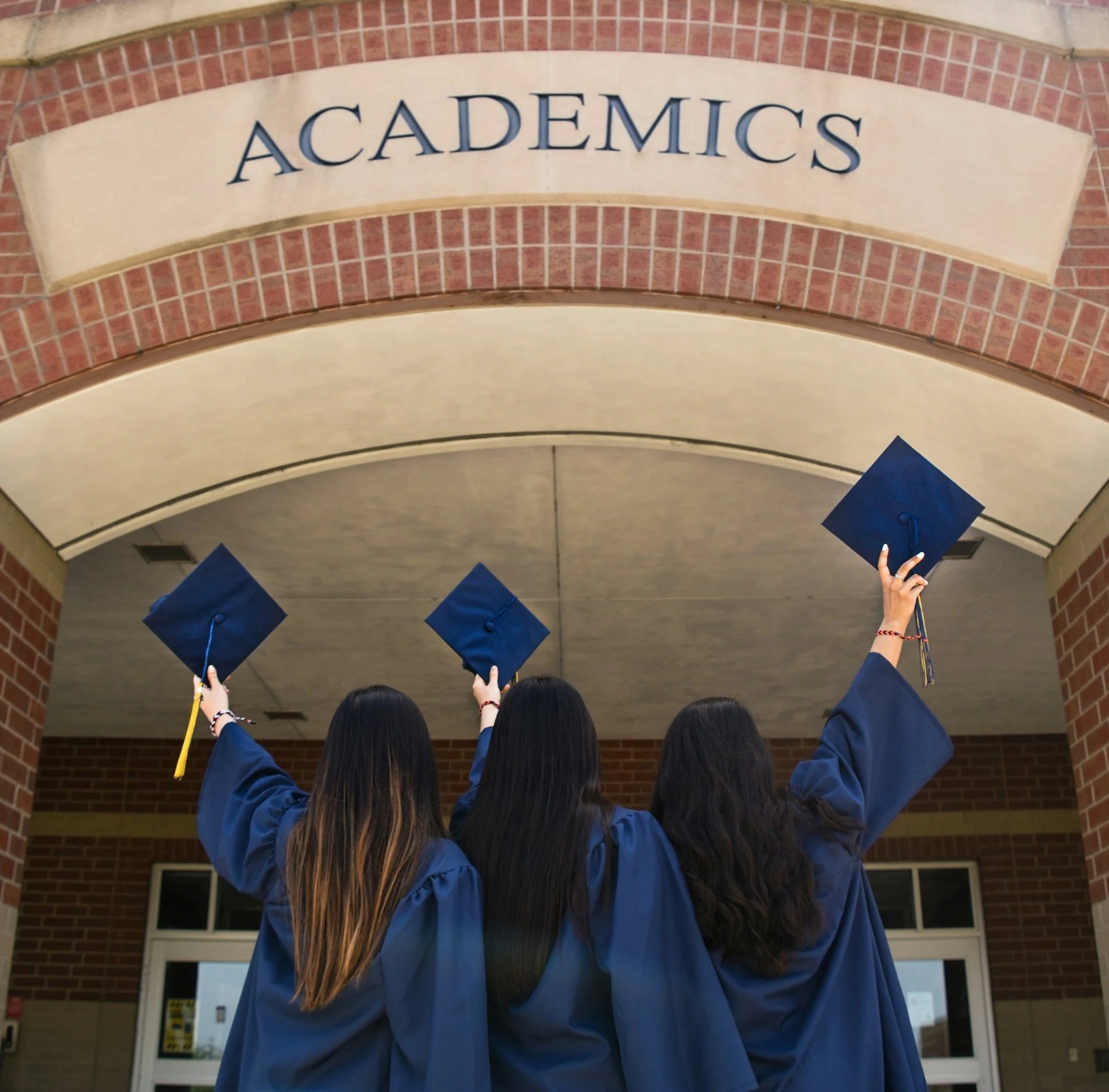 Students in blue gowns holding diplomas celebrating graduation outside a brick building with an arched entrance.