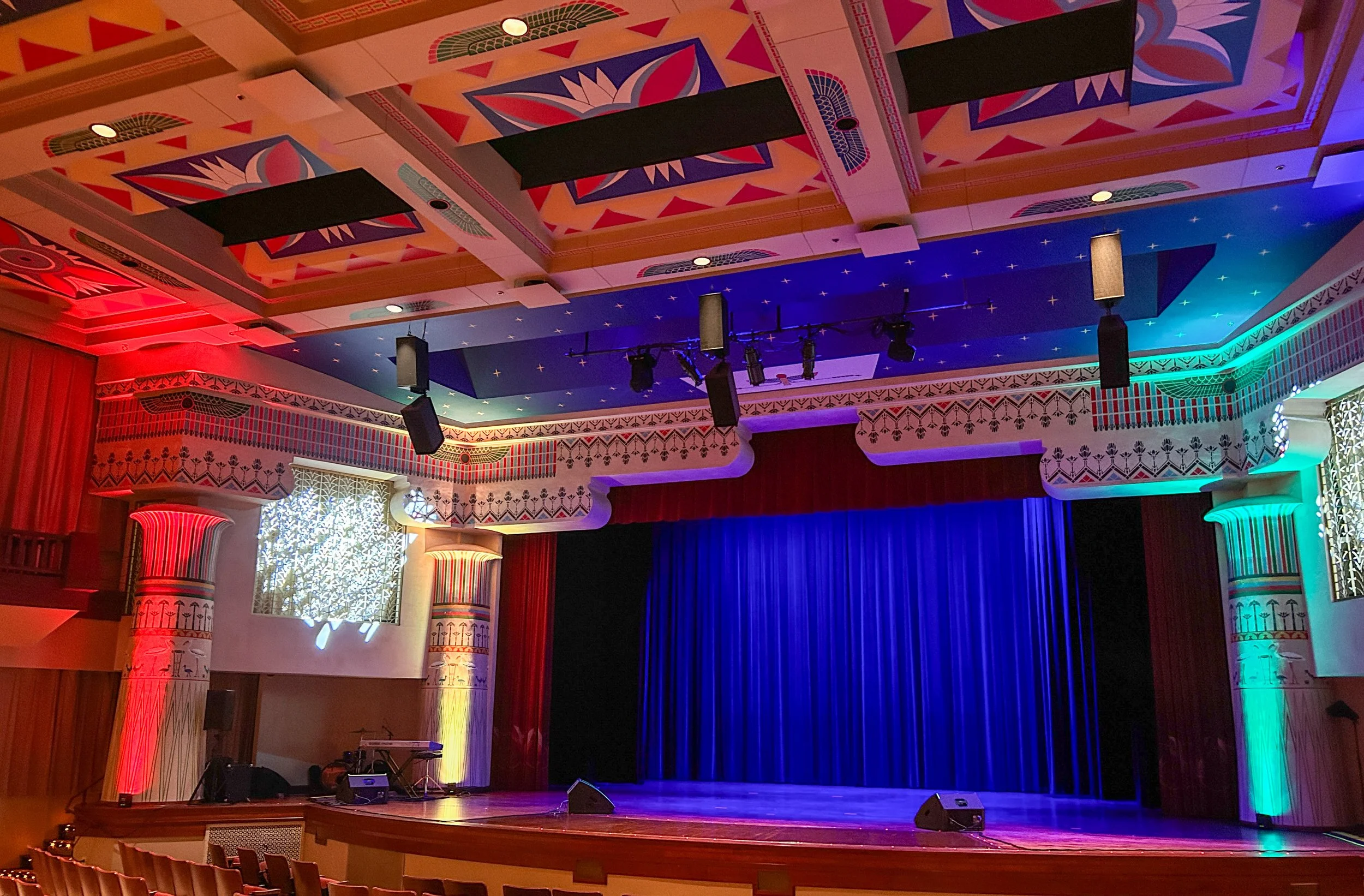 Interior of a theater or auditorium with a decorated ceiling, stage with red curtains, and colorful lighting.