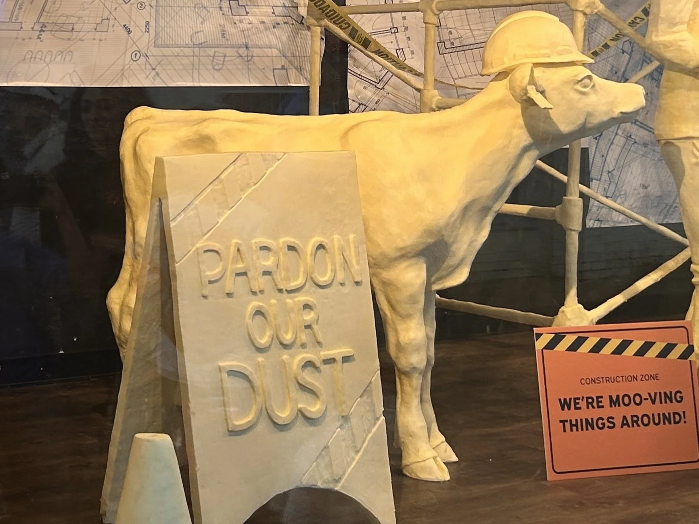 A sculpture of a dog wearing a construction helmet, standing next to a sign that reads 'Pardon Our Dust'. There is a construction zone sign nearby stating, 'We're Moo-ving Things Around!'. The background features construction drawings and scaffolding.