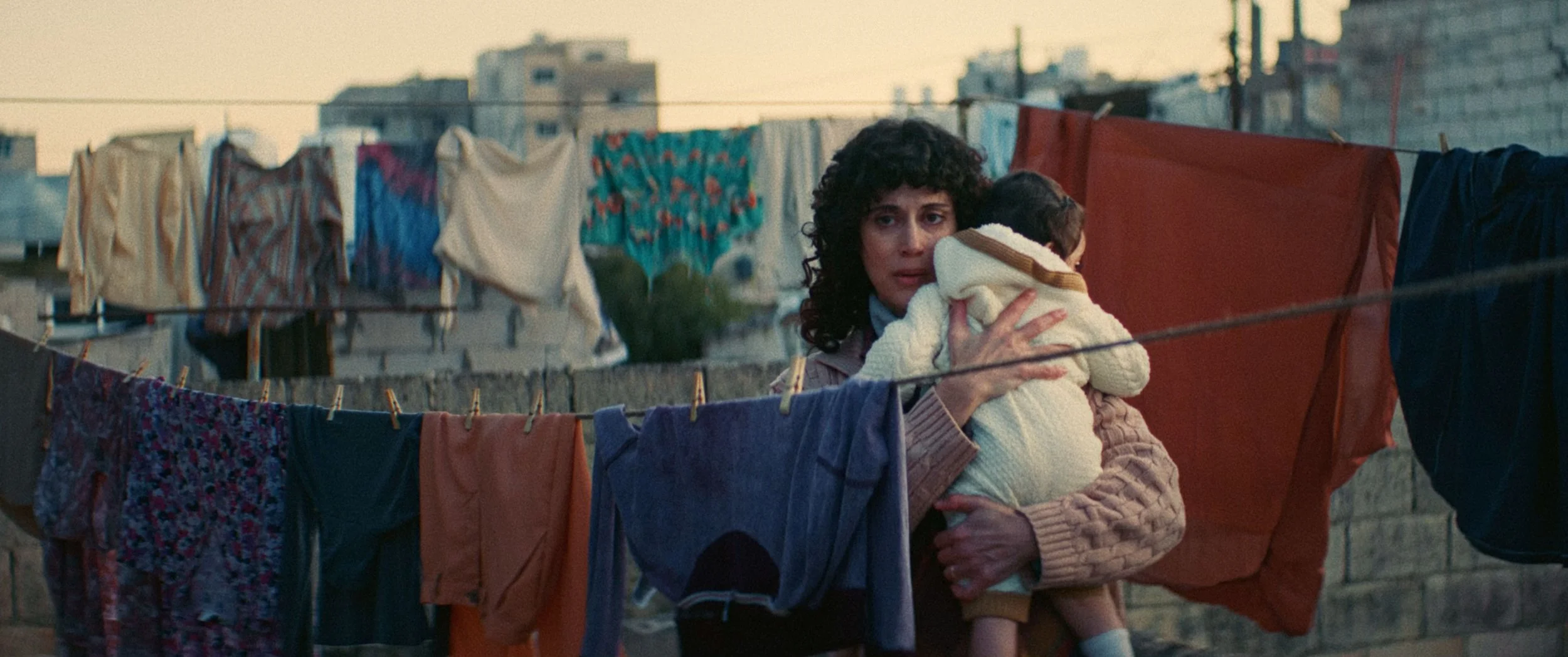 A woman holding a child while standing among hanging laundry on clotheslines in an urban neighborhood.