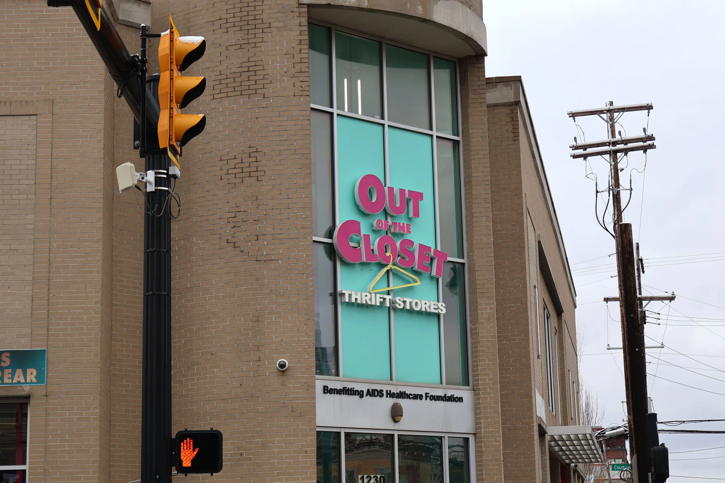A corner of a brick building with a large vertical sign that reads 'OUT OF THE CLOSET' in pink letters and 'THRIFT STORES' in white letters, indicating a thrift store benefiting AIDS Healthcare Foundation. A traffic light showing red is visible in the foreground, and there are electrical poles and wires nearby.