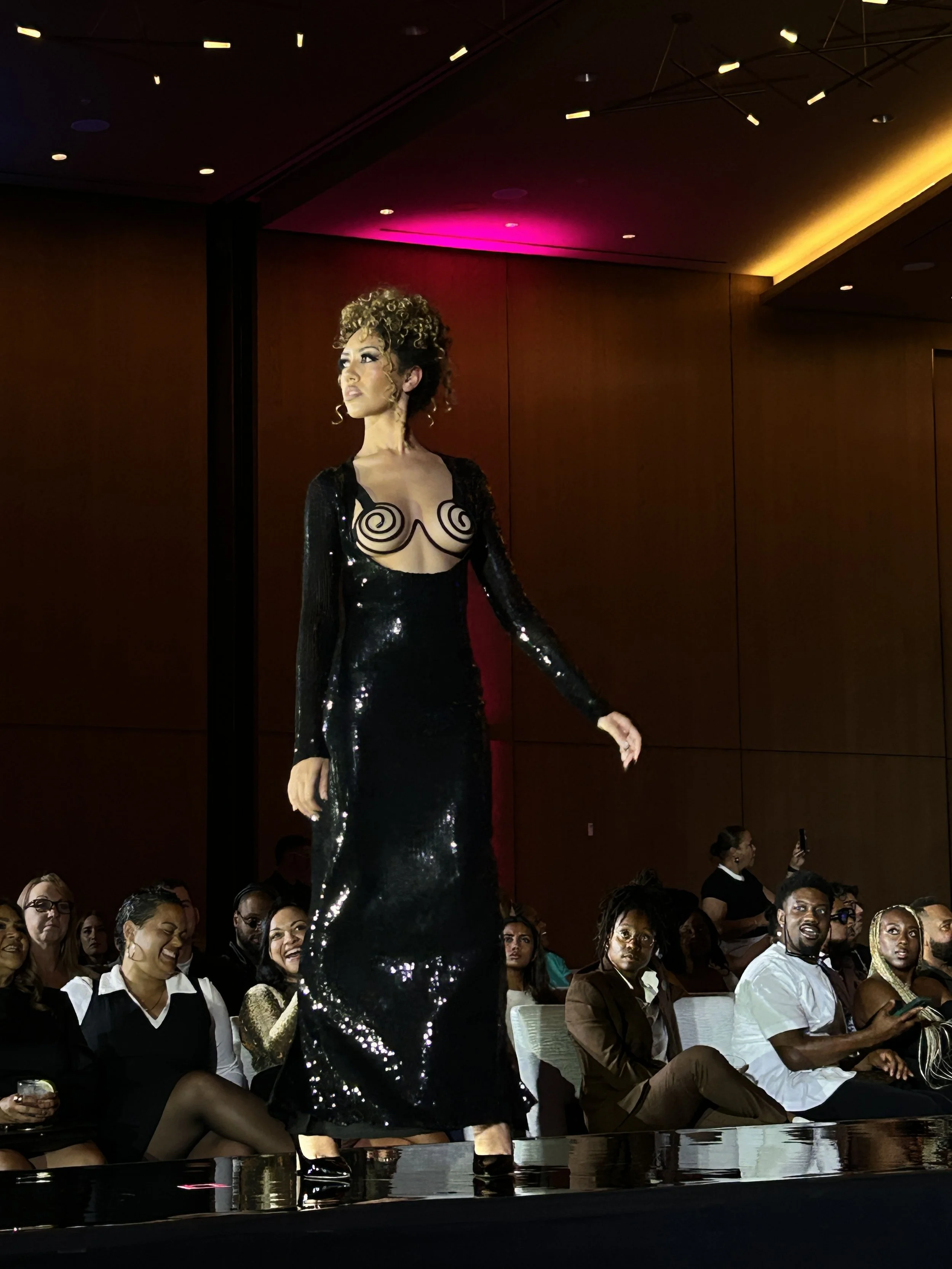 A woman in a black sequined dress with spiral patterns on her chest stands confidently on a stage at an event, with an audience seated below, some engaged and others taking photos.