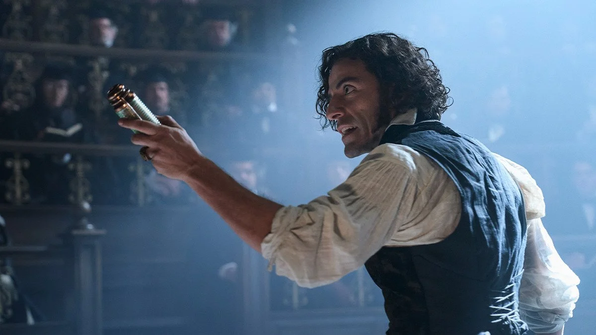 A man with dark curly hair, wearing a beige shirt and a black vest, holding a weapon and pointing it aggressively in a scene with an audience in the background.