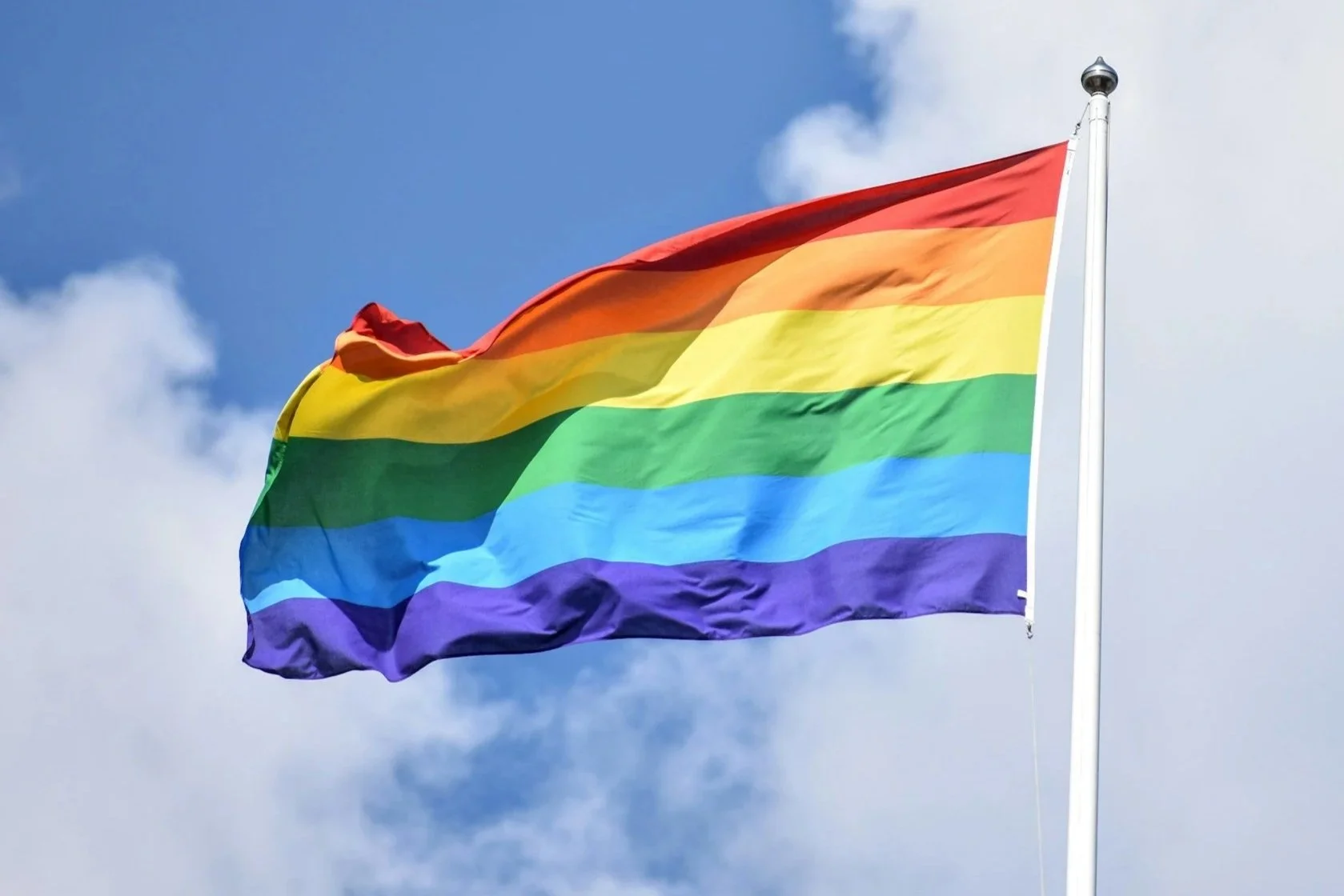 Rainbow pride flag flying outdoors against a partly cloudy sky.