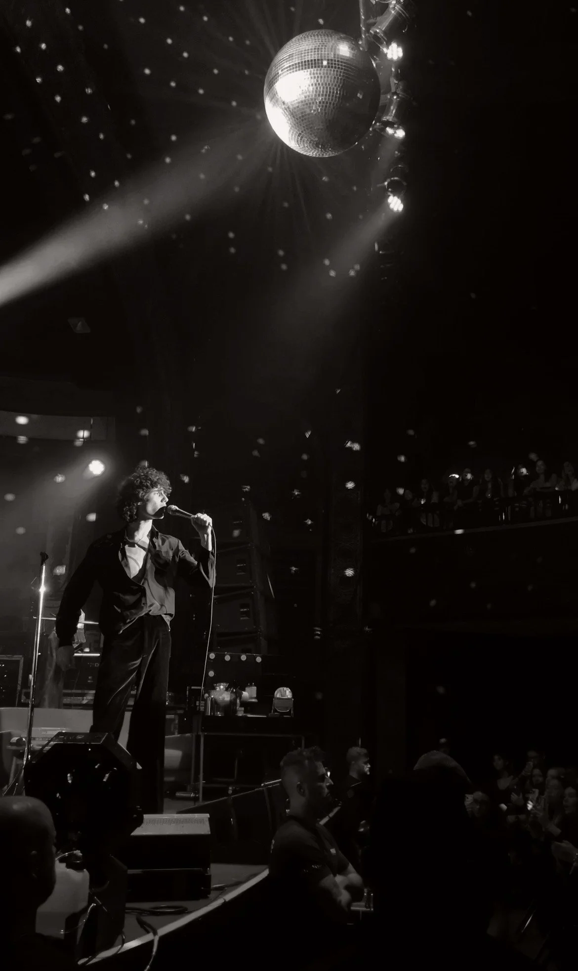 A performer with curly hair sings into a microphone on stage under a disco ball with lights and audience members watching from both the stage and balcony.