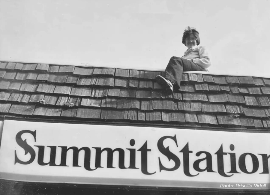 A child sitting on a tiled roof above a sign that reads 'Summit Station' with a photo credit to Priscilla Ridal.