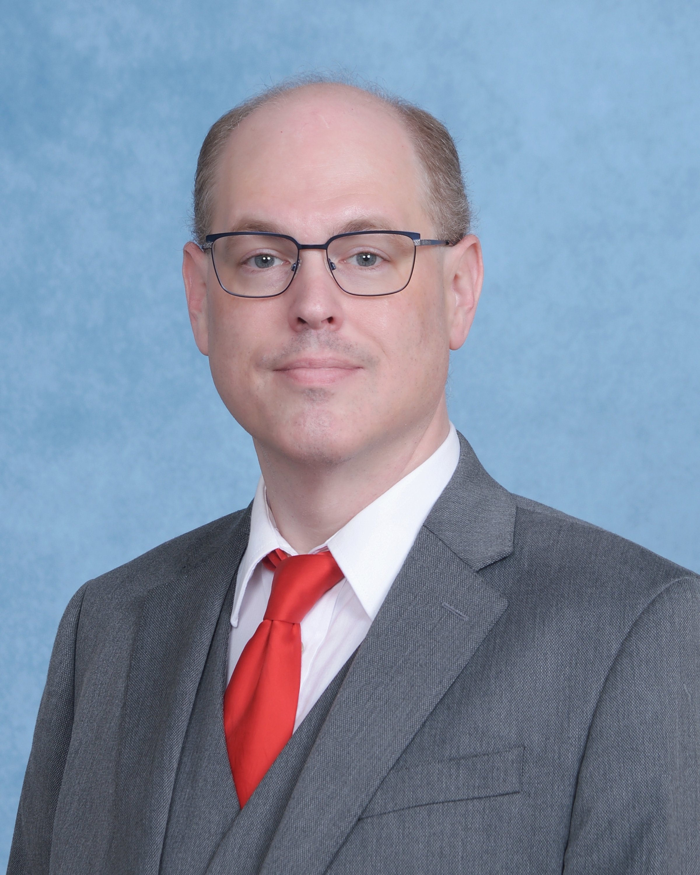 A man wearing a gray suit, white shirt, red tie, and glasses, with a blue background.