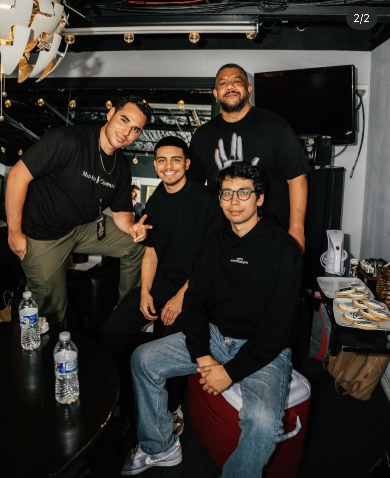 Four young men posing together for a photo indoors, with a table of donuts and bottled water nearby, and a television mounted on the wall behind them.