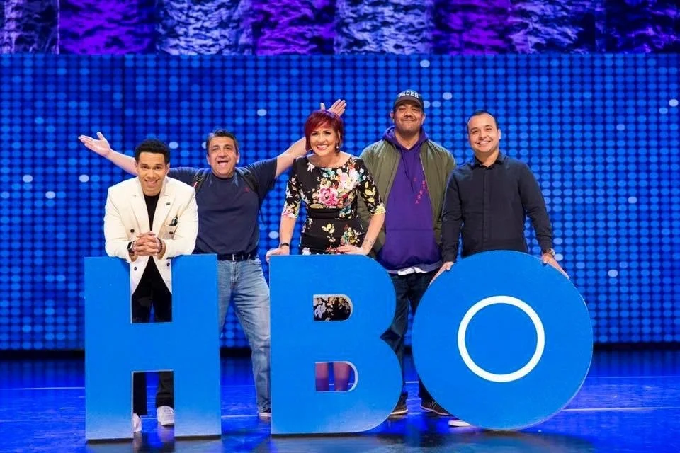 Five people standing behind large blue letters spelling 'HBO' on a stage with a blue lit backdrop.