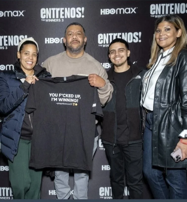 Four people standing together in front of a backdrop with logos for HBO Max and Entrelos, holding a black T-shirt that reads 'You f*cked up, I'm winning!' in white text.