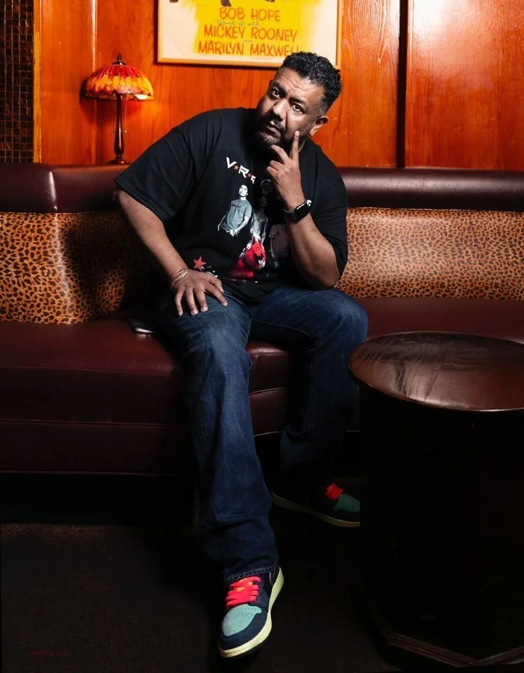 A man sitting on a leather booth, with a wooden wall and a lamp in the background, and a framed poster above him. He is wearing a black graphic T-shirt, jeans, and colorful sneakers, and is holding his chin with one hand while looking at the camera.