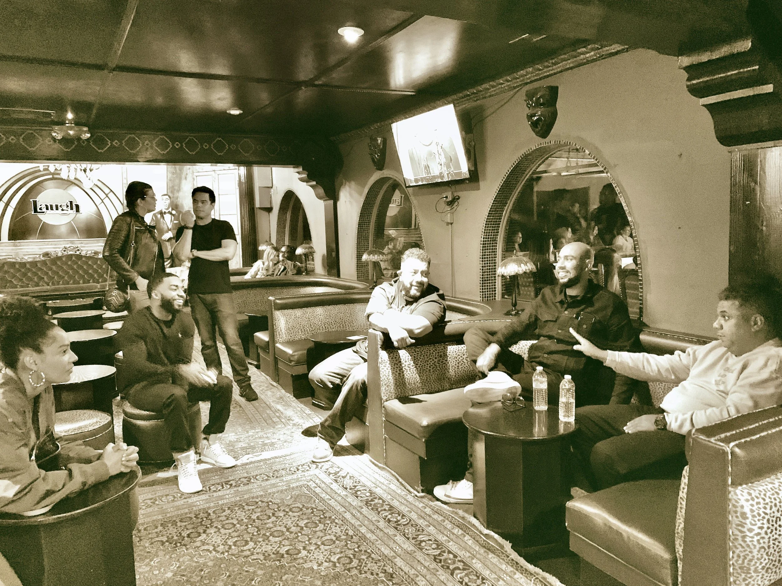 A group of people sitting and standing in a cozy, vintage-style bar or lounge. Some are engaged in conversation, others are smiling or looking around. The room features decorative arches, a patterned carpet, and dim lighting.