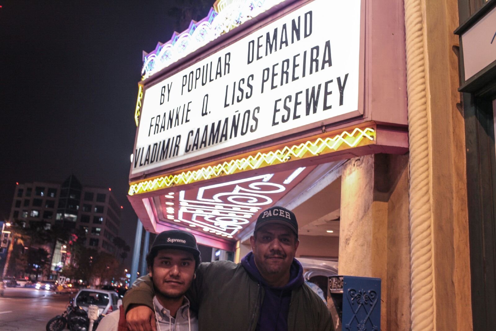 Two men standing in front of a theater marquee at night. The marquee displays the names of performers Frankie Q. Liss Pereira and Vladimir Caamano Esewey. The men are smiling, wearing hats, one with a Supreme cap and the other with a PACE cap. The ba