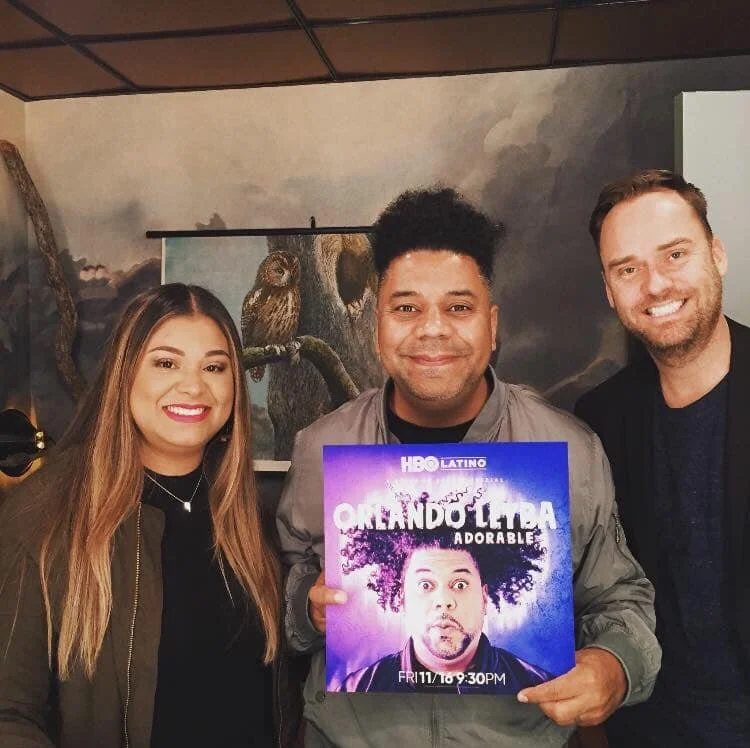 Three people smiling and holding a promotional sign for the HBO Latino show 'Oregando La Vida Adorable.' The man in the center is displaying the sign, which features a photo of a man with curly hair and a surprised expression, with the show's title a