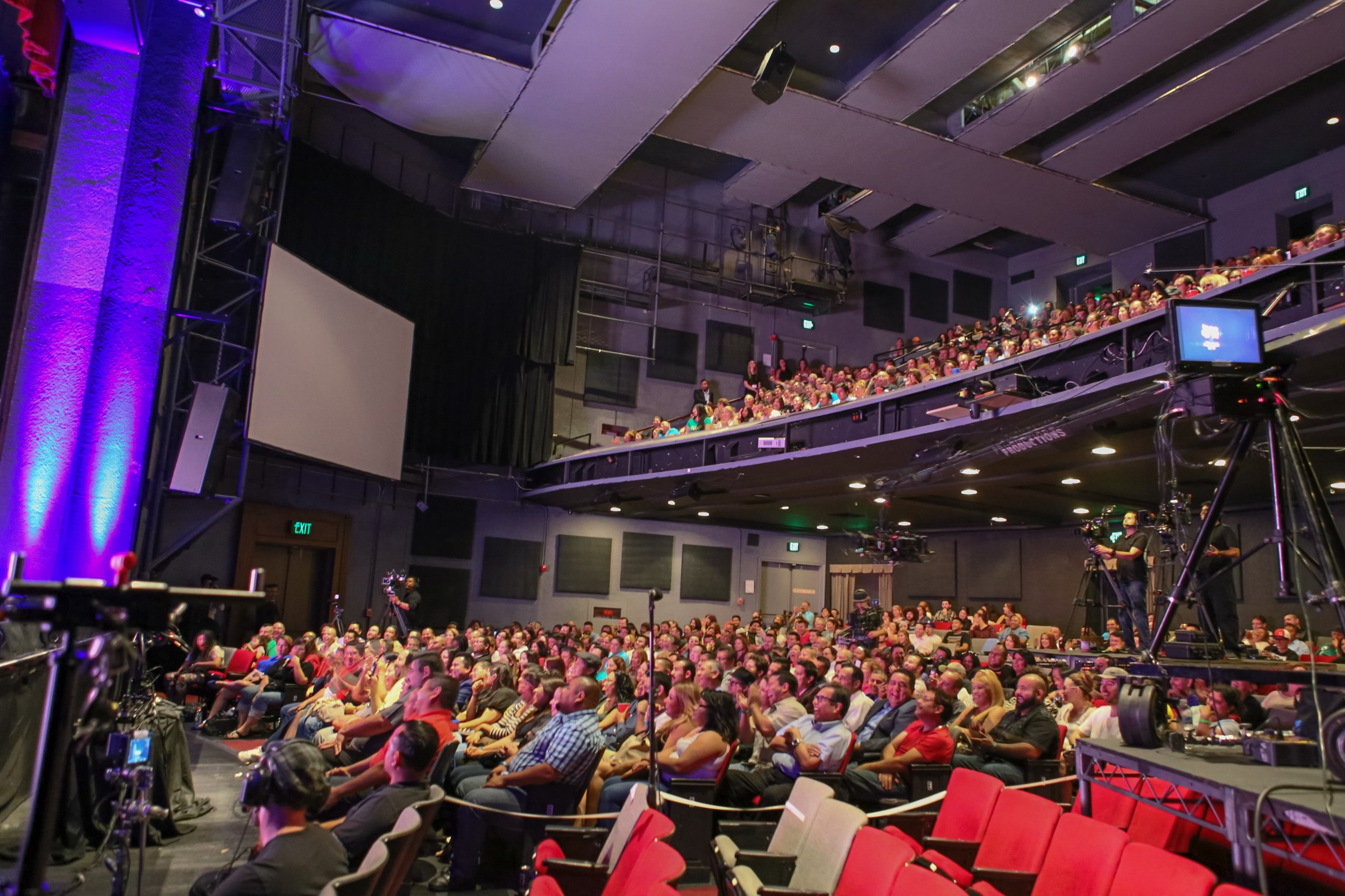 Audience seated in a theater or auditorium, facing a stage, with some people standing behind a railing on the balcony level. The setting is dimly lit with stage lighting, and a large projection screen is visible on the left side of the stage. The cro