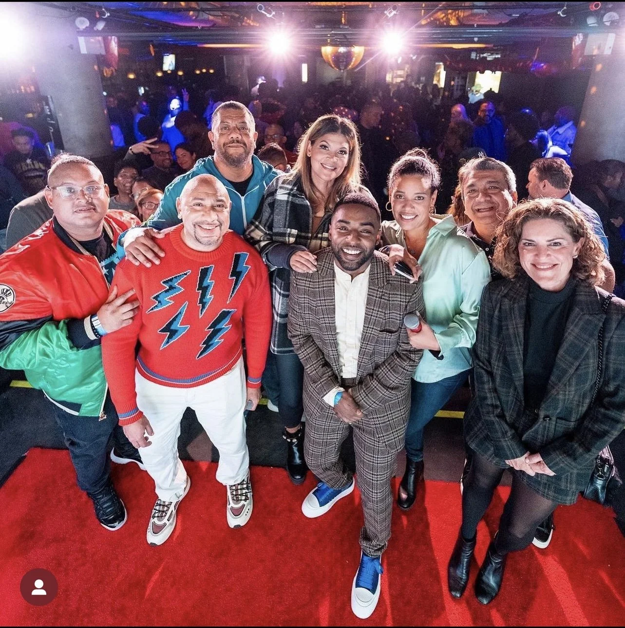 Group of diverse people smiling and posing for a photo on a red carpet at a lively indoor event with a crowded background and colorful lighting.