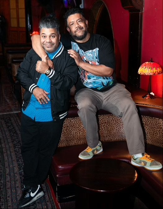 Two men posing together in a cozy, dimly lit room with red walls. One man is standing, giving a thumbs-up and wearing a black jacket over a blue shirt, while the other is sitting on a booth seat with his arm around the standing man, wearing a black T