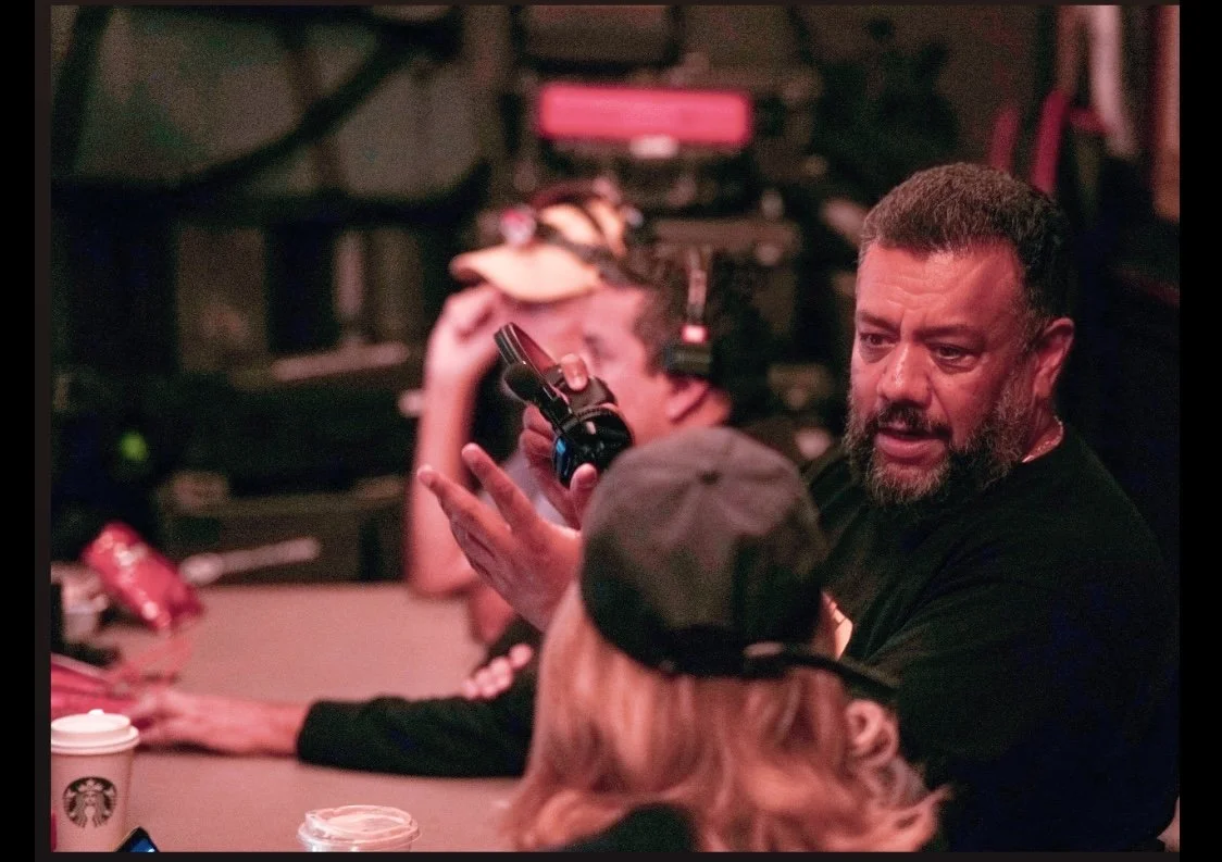 A man with a beard and short hair speaking while gesturing with his hands at a group gathered around a table in a dimly lit room, possibly a bar or cafe, with coffee cups on the table and blurred background featuring electronic equipment and a person